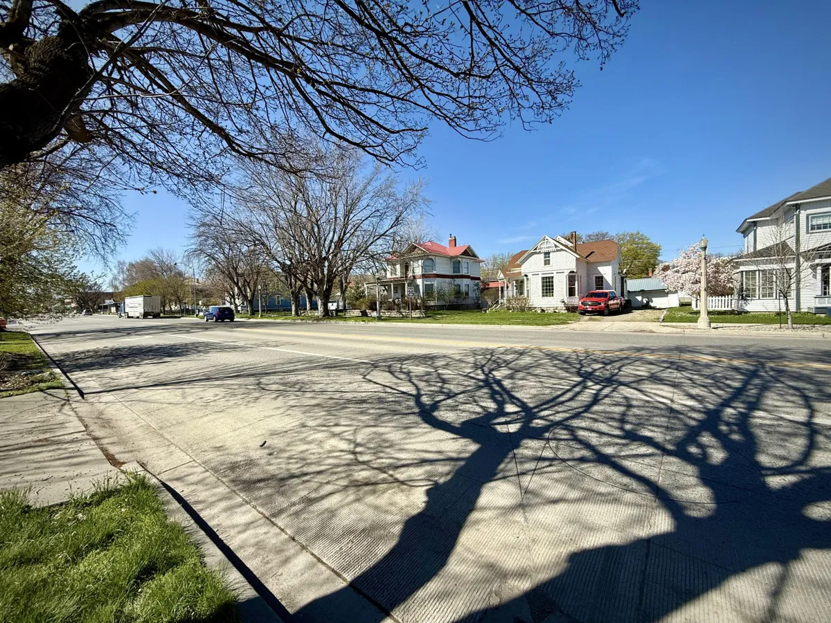 A tranquil neighborhood street bordered by houses of varied architecture, illuminated by tree shadows and set beneath a clear blue sky. The scene captures the welcoming, walkable environment of Baker City's residential area.