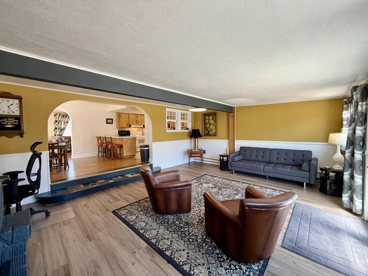A charming living area featuring a combination of armchairs and a sofa around a decorative rug, with an arched doorway offering a glimpse toward the adjoining kitchen. The comfortable seating and inviting color palette create a homey feel, while the open layout underlines the home's flow and connectivity.