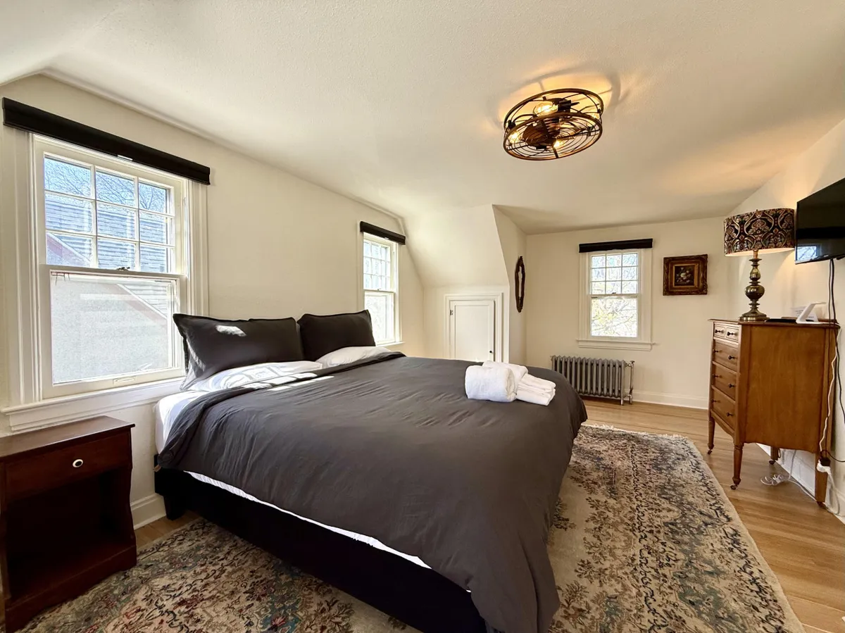 A spacious bedroom with a king-sized bed dressed in dark bedding, windows letting in abundant natural light, a decorative dresser, and a modern ceiling fixture. The overall feel is calm, serene, and ideal for both quiet evenings and gentle morning awakenings.