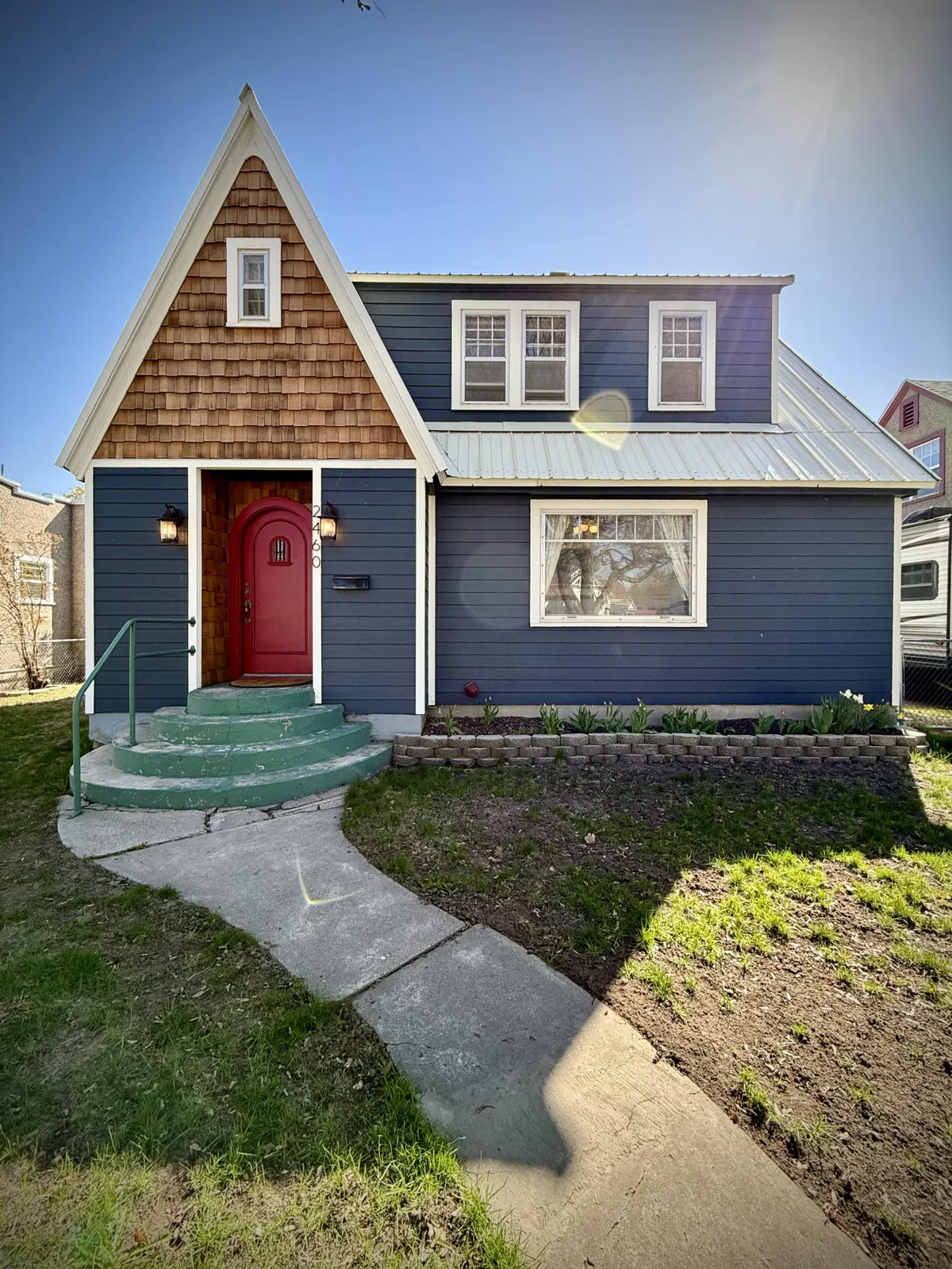 A welcoming view of Storybook Nook's exterior, featuring a red front door, classic shingle roof, blue siding, and inviting steps leading up to the entrance. The front yard hints at the possibility of gatherings and seasonal activities.