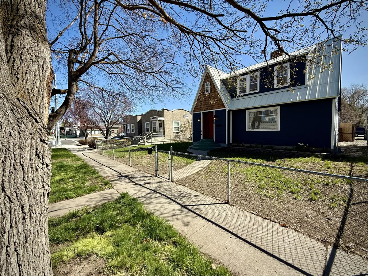 A well-maintained blue house with a distinctive roof and a tidy front yard featuring a prominent tree. The sidewalk and open yard suggest a lively, friendly neighborhood perfect for afternoon strolls or community gatherings.