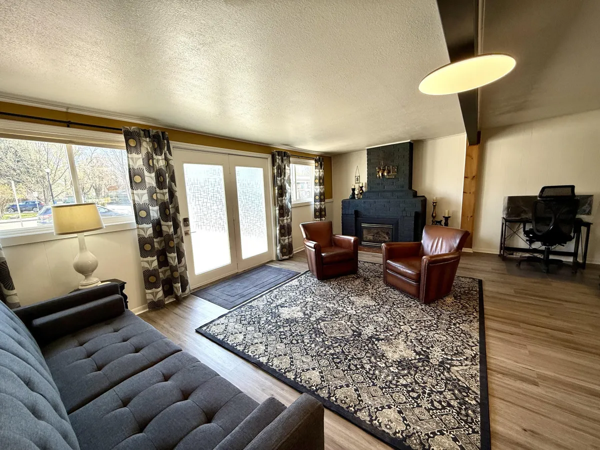 A cozy living room with a glowing fireplace as the centerpiece, surrounded by a plush sofa, two comfortable armchairs, a patterned area rug, and large windows that fill the space with natural light. Eclectic artwork and decor add personality and warmth to the scene, creating an inviting setting for gatherings or quiet reading.