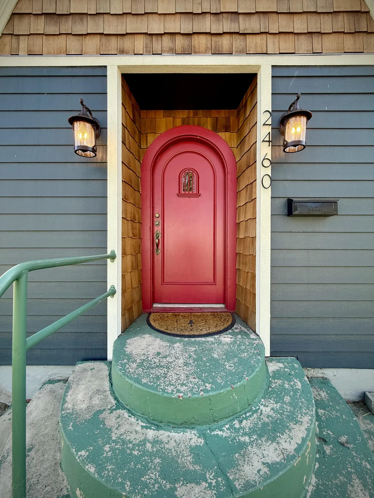 The inviting red front door of a home framed by an arched doorway, decorative light fixtures, and green circular steps. The textured wooden shingles and festive touches suggest a warm, welcoming entrance typical of Baker City's historic district.