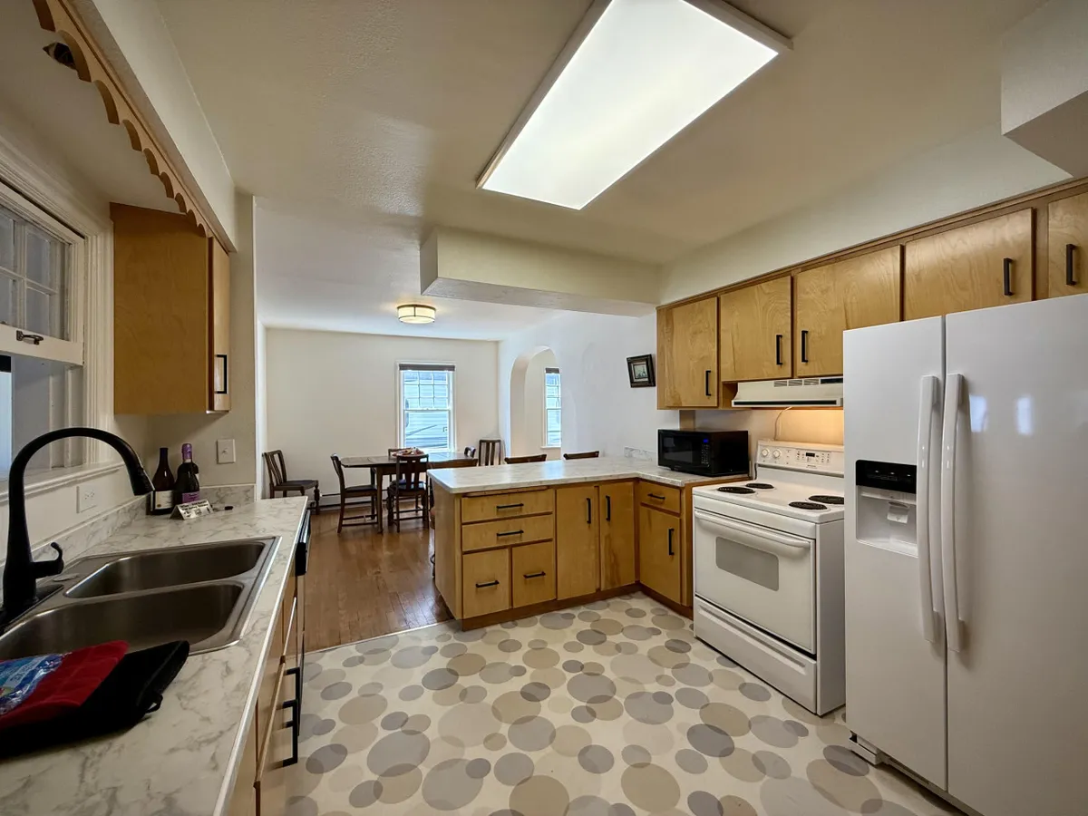 A cozy kitchen and dining area featuring a kitchen sink, stove, refrigerator, and a visible dining table. The scene is suffused with warm, natural light, embodying a sense of comfort and readiness for shared meals or coffee breaks.
