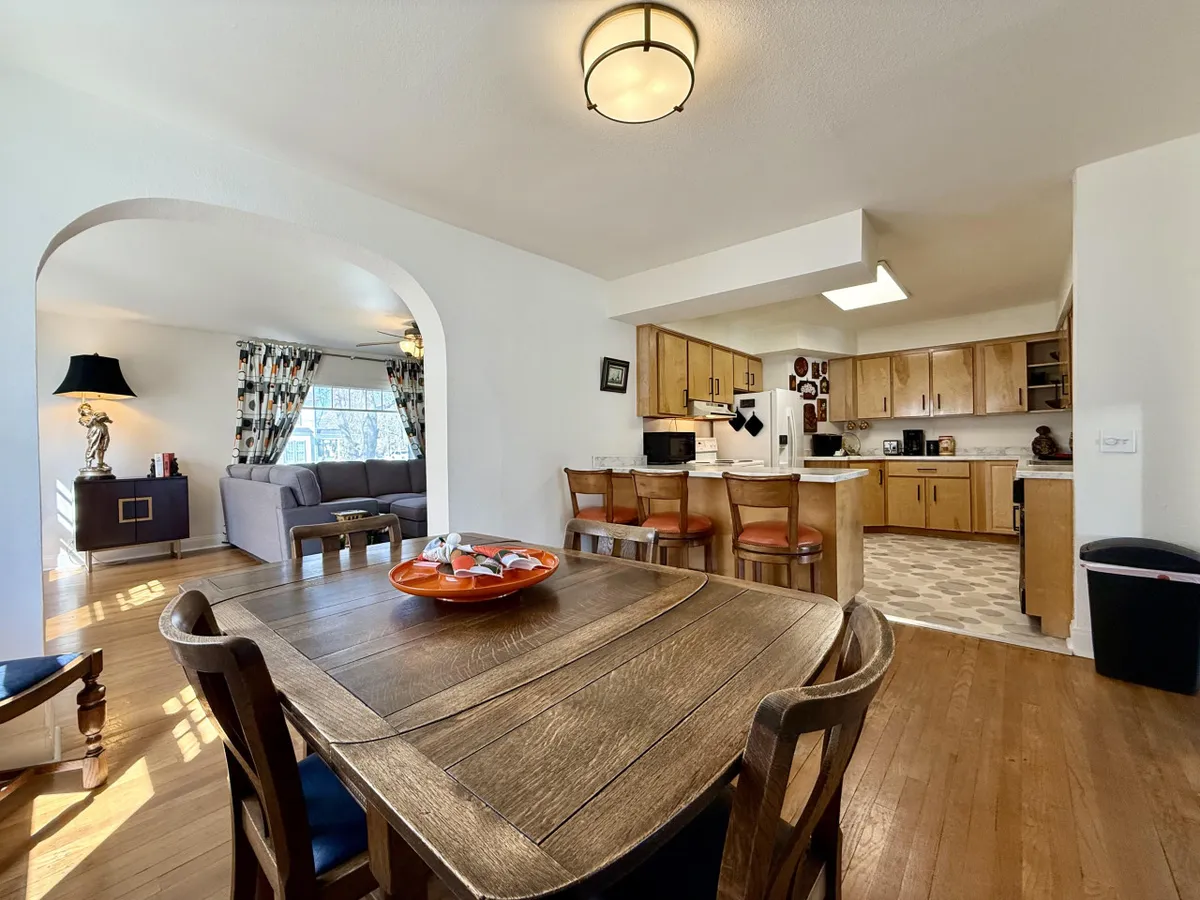 A dining table set for six with an orange bowl centerpiece, an archway leading to the living area, and the kitchen visible in the background. The open layout visually connects kitchen, dining, and living spaces, highlighting both the seating capacity and the home’s sociable design.